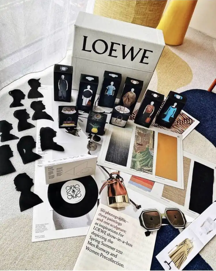 L’ensemble d’archive « Show in a Box » LOEWE mis en scène sur un tapis, avec les silhouettes de l’équipe, les boîtes de vêtements miniatures, le disque vinyle et les lunettes de soleil
